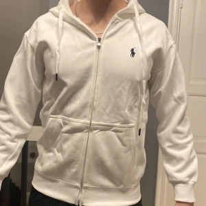 Vit hoodie från Polo Ralph Lauren - Säljer en stilren vit hoodie från Polo Ralph Lauren med dragkedja och det klassiska logotypen på bröstet. Tröjan har en bekväm passform med långa ärmar och en praktisk ficka framtill. Perfekt för en avslappnad look.