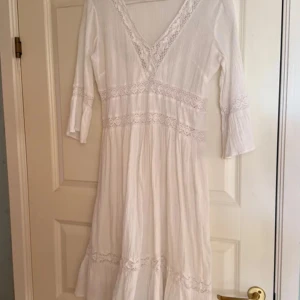   studentklänning  - Charmig vit klänning med v-ringning och spetsdetaljer. Klänningen har trekvartsärmar och en luftig passform som ger en romantisk känsla. Perfekt för sommarens alla tillfällen. Bomull med stretch. Fodrad .
