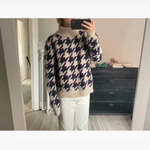 Beige mönstrad stickad polotröja - Säljer en mysig beige stickad polotröja med ett mönster i blått och rött. Tröjan har långa ärmar och en hög polokrage som håller dig varm under kyliga dagar.