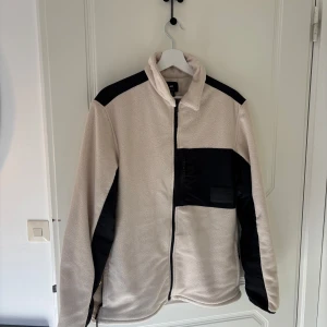 Beige fleecejacka från H&M - Säljer en beige fleecejacka från H&M med svarta detaljer. Jackan har en dragkedja framtill och en hög krage. Perfekt för vår och höst.