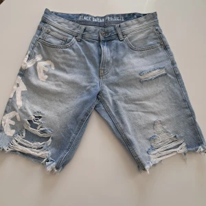 Jeansshorts från Black Squad Projects - Snygga ljusblå jeansshorts med slitna detaljer och fransiga kanter. De har trycket 'LIVE OR DIE' i vitt på baksidan och flera fickor. Perfekta för en avslappnad stil.