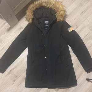 Svart parkajacka från Svea - Säljer en svart parkajacka från Svea med en mysig pälsfodrad huva. Jackan har en amerikansk flagga på ärmen och stängs med dragkedja och knappar. Perfekt för kalla dagar!