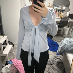 Ljusblå knytblus från Gina Tricot - Säljer en stilren ljusblå blus från Gina Tricot med knytning framtill. Blusen har långa ärmar och en v-ringad design som ger en elegant touch. Perfekt för att lyfta vilken outfit som helst.