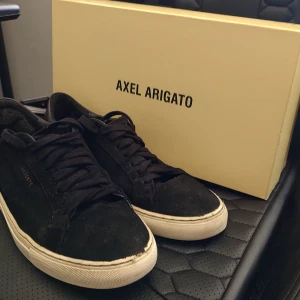 Svarta sneakers från Axel Arigato - Snygga svarta sneakers från Axel Arigato i mocka med vit sula. Skorna har klassisk snörning och en stilren design som passar perfekt till vardagsoutfiten. Perfekta för dig som gillar en minimalistisk stil. Slutet framtill på högra skon. Samt att "arigato" försvunnit nästan helt på den vänstra skon. 