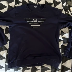 Mörkblå tröja från Emporio Armani - Står xxl men sitter som en xl/L.  .Säljer en stilren mörkblå tröja från Emporio Armani med det ikoniska EA7-logotypen på bröstet. Tröjan har långa ärmar och en rund halsringning. Perfekt för en avslappnad och trendig look. 