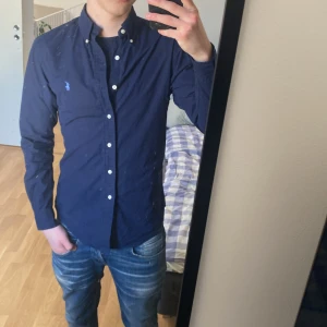 Mörkblå skjorta från Ralph Lauren - Säljer en stilren mörkblå skjorta från Ralph Lauren i slim fit. Skjortan är tillverkad i 100% bomull med stretch och har knappar framtill samt en broderad logga på bröstet. Perfekt för en snygg och avslappnad look.