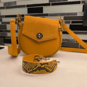 Gul axelväska med ormmönstrade detaljer - Snygg gul axelväska med unika ormmönstrade detaljer på sidorna och axelremmen. Väskan har en rund form och stängs med ett metallspänne framtill. Perfekt för att ge en färgklick till din outfit!