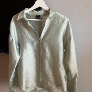 Glittrig ljusgrön skjorta Gina Tricot - Superfin glittrig skjorta från Gina Tricot i ljusgrönt med skimrande yta. Skjortan har klassisk krage, knappar framtill och lång ärm. Litet hål (se sista bild) som man inte märker av.