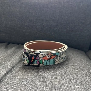 Louis Vuitton bälte med tecknad print - Louis Vuitton bälte i skinn med färgglatt tecknat mönster och svart LV-spänne. Bältet har inslag av blått, vitt och orange samt tydlig logga framtill. Insidan är ljusbrun och slät med präglad märkning. Perfekt för dig som vill sticka ut med en unik accessoar.