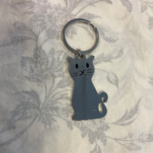 Nyckelring med katt i silverfärg - Söt nyckelring i form av en katt, tillverkad i metall med blank silverfärg och svarta detaljer för ögon, nos och morrhår. Katten har en stilren och lekfull design, perfekt att hänga på väskan eller nyckelknippan.