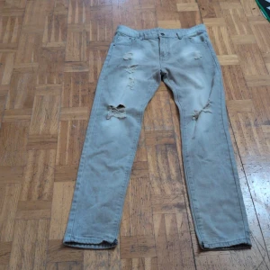 Ljusgrå ripped jeans 14-15 år - Säljer ett par ljusgrå jeans med slitna detaljer och hål på benen. Jeansen har klassisk femficksdesign och är tillverkade i bomull och polyester. Perfekta för en avslappnad och trendig look.