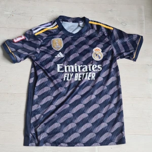 Real Madrid bortaställ Adidas blå - Säljer ett Real Madrid bortaställ från Adidas. Tröjan är marinblå med vågigt mönster, gula detaljer på axlarna och klubbmärke samt sponsortryck framtill. Matchande shorts med gula och vita ränder på sidorna. Shortsen ser ut att vara små i midjan men det är dem inte utan de är bara väldigt stretchiga. Tillverkat i lätt och ventilerande polyester. Har typ aldrig använt då jag är ett Barca fan och fick detta i födelsedagspresent. Kan sälja shortsen för 100 och tröjan för 150 separat.
