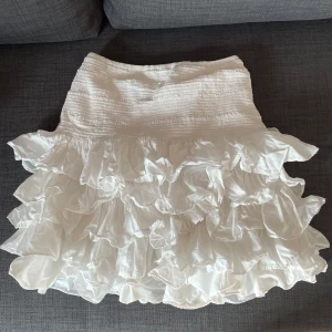 Vit volangkjol från H&M, storlek S - Supersöt vit kortkjol från H&M med flera lager volanger och smockad midja. Kjolen är i tunn bomull och har en luftig känsla som passar perfekt till varma dagar. Enkel att styla med crop top eller oversized t-shirt för en chill vibe.