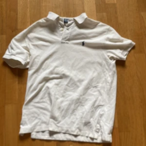 Vit pikétröja från Polo Ralph Lauren - Klassisk vit pikétröja från Polo Ralph Lauren i bomull. Tröjan har korta ärmar, krage och knäppning framtill samt den ikoniska blå broderade loggan på bröstet. Perfekt för en clean och stilren look.