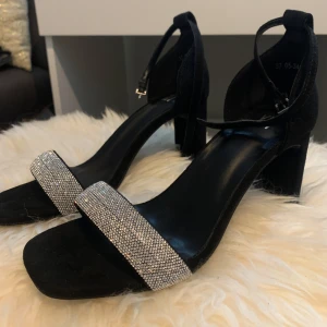 Svarta klackskor med strass från XIT - Snygga svarta klackskor från XIT med bred rem framtill täckt av glittrande strass. Skorna har öppen tå, blockklack och smal ankelrem med spänne. Perfekta för att sticka ut med en elegant touch och bekväm passform.
