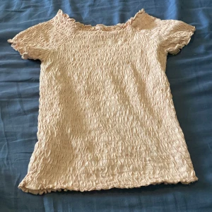 Beige smockad topp från H&M - Supersöt smockad topp från H&M i ljusbeige med broderad text 'KINDNESS' framtill. Toppen har korta ärmar, vågig kant och är gjord i mjuk bomull. Perfekt för dig som gillar en chill och trendig stil.