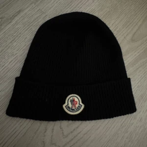 Svart ribbad mössa från Moncler - Svart ribbstickad mössa från Moncler med klassisk uppvikt kant och broderad Moncler-logga framtill. Tillverkad i mjukt material som håller dig varm och ger en clean look. Perfekt accessoar för kalla dagar och enkel att matcha med olika outfits. Kvitto finns 