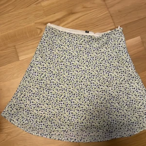 Blommig kortkjol från Divided H&M - Supersöt kortkjol från Divided H&M i vitt med små blå blommor och gröna blad. Kjolen har en lätt A-linjeform och är gjord i ett mjukt, tunt material som känns perfekt för varma dagar. Perfekt att styla med en enkel topp och sneakers.