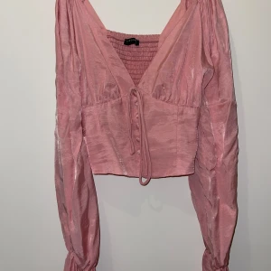 Topp - Supersöt rosa omlottopp. Toppen har långa puffiga ärmar, v-ringning, knyt framtill och klädda knappar. Smockad rygg för extra komfort och croppad passform. Perfekt för dig som gillar romantiska detaljer och trendig stil.
