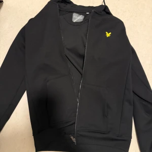 Svart hoodie från Lyle & Scott - Svart hoodie från Lyle & Scott med gul logga på bröstet. Tröjan har huva, dragkedja framtill och två stora fickor. Perfekt för en chill och stilren look. Materialet är mjuk bomull som känns skönt mot huden.