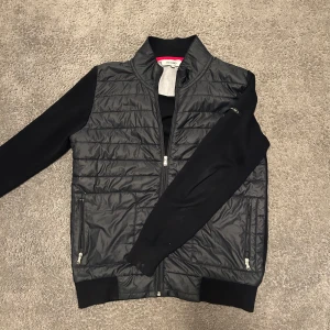 Cardigan från Iceland - Svart/mörkblå cardigan från Iceland med glansig framsida och ärmar i mjukare material. Jackan har dragkedja framtill och två fickor med dragkedja. Perfekt för kyliga dagar när du vill ha en stilren look. Funkar super till svalare sommarkvällar!! 