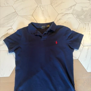 Marinblå pikétröja från Polo Ralph Lauren - Klassisk marinblå pikétröja från Polo Ralph Lauren med korta ärmar och röd broderad logga på bröstet. Tröjan har krage och knappar vid halsen, tillverkad i mjuk bomull som känns skön mot huden.