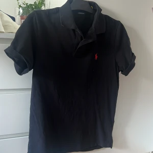 Svart pikétröja Polo Ralph Lauren - Svart pikétröja från Polo Ralph Lauren i custom slim fit. Klassisk krage, korta ärmar och röd broderad logga på bröstet. Tillverkad i mjuk bomull, perfekt för en clean och stilren look.