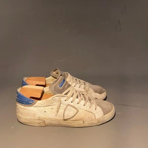 Philippe Model sneakers beige/blå mocka - Sneakers från Philippe Model i beige mocka och skinn med blå detaljer på hälen. Klassisk låg modell med rund tå, snörning och platt sula. Märkeslogga på sidan och tungan. Perfekta för en avslappnad och trendig look.