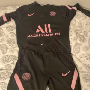 PSG svart och rosa träningsset Nike - Snyggt svart träningsset från PSG med rosa detaljer och loggor. Tröjan har lång ärm, halv dragkedja och tryck med Accor Live Limitless. Byxorna har elastisk midja och rosa ränder längs benen. Perfekt för dig som älskar fotboll och vill sticka ut.