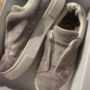 Grå mockasneakers från Eytys - Snygga sneakers från Eytys i ljusgrå mocka med minimalistisk design och vit platt sula. Skorna har rund tå och elastisk plös utan snörning för en clean look. Perfekta för dig som gillar stilrena och trendiga sneakers med premiumkänsla.