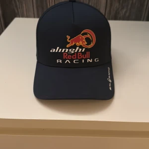 Alinghi Red Bull Racing keps Sail Racing - Mörkblå keps från Sail Racing med Alinghi Red Bull Racing-logga framtill och Tudor-logga på sidan. Justerbar rem baktill med metallspänne och schweiziskt kors. Tillverkad i 100% polyester, perfekt för dig som gillar racing och snygga accessoarer.använd 2 gånger 