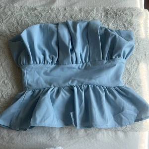 Ljusblå volang tubtopp - Supersöt ljusblå tubtopp med bred volang nertill och snygga veck upptill. Toppen är axelbandslös och har dragkedja i ryggen. Perfekt för dig som vill sticka ut med en feminin och trendig look. Storlek S men är ganska stor i storleken så skulle snarare säga att det är mer som en M. Helt ny och aldrig använd!!🫶