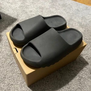 Adidas Yeezy Slide i mörkgrå - Adidas Yeezy Slide i mörkgrå färg med chunky, räfflad sula och öppen tå. Tillverkade i syntetmaterial för en clean och futuristisk look. Perfekta för chill dagar och streetwear-stil. Storlek 47(sitter som en 46)