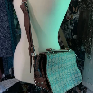 Brun axelväska med dödskallemönster - Cool brun axelväska i skinnimitation med all-over dödskallemönster och silverfärgade nitar på sidorna. Justerbar axelrem och lockstängning. 