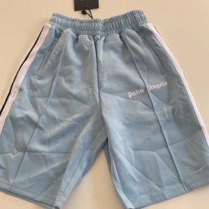 Palm Angels ljusblå shorts  - Ljusblå shorts från Palm Angels med vita och svarta revärer längs sidorna. Elastisk midja och broderad logga framtill. Sportig vibe och tillverkade i ett mjukt polyestertyg som känns skönt mot huden. Perfekta för sommaren och chill dagar.