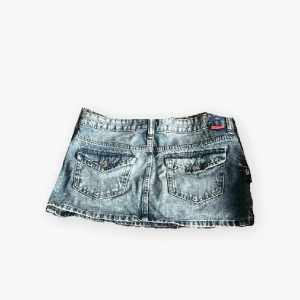 Blå jeanskjol med fickor - Snygg kort jeanskjol i klassisk blå denim med slitna detaljer. Kjolen har två bakfickor med lock och knappar, samt bälteshällor och knappstängning fram. Perfekt för dig som gillar en avslappnad och trendig stil.