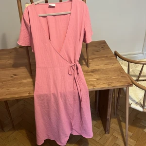 Rosa omlottklänning från Vila - Superfin rosa omlottklänning från Vila i storlek 36. Klänningen har korta ärmar, knytband i midjan och v-ringad hals. Materialet är mjukt och lätt, perfekt för varma dagar. Enkel och stilren design som känns både trendig och bekväm.