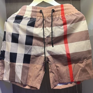 Rutig Burberry badshorts med snörning - Snygga badshorts från Burberry med klassiskt rutigt mönster i beige, svart, vitt och en röd rand. Shortsen har elastisk midja med svart snörning och innerfoder i mesh. Perfekta för stranden eller poolen om du vill sticka ut med stil. Pris går alltid att diskutera 