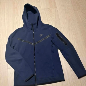 Mörkblå Nike tech träja med mjukisbyxor med dragkedja - Snygg mörkblå tröja med mjukisbyxor från Nike  och dragkedjeficka på benet. Byxorna är tillverkade i mjukt material och har en sportig look som passar perfekt till träning eller chill. Klassisk Nike-logga och stilren design.