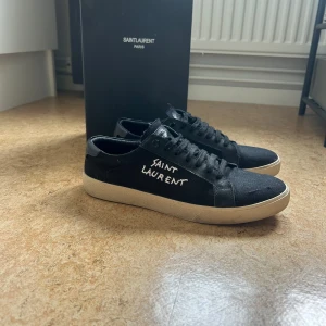Svarta Saint Laurent sneakers - Svarta sneakers från Saint Laurent med vit text på sidan och klassisk låg siluett. Ovandel i canvas och detaljer i skinn, svarta snören och vit platt sula. Snygg minimalistisk design med diskret logga på plösen och insidan.