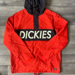 Röd vindjacka från Dickies med huva - Snygg röd vindjacka från Dickies med svart huva och stor logga över bröstet. Jackan har lång ärm, dragkedja vid halsen och justerbar nederkant med snörning. Perfekt för dig som vill sticka ut med streetstyle-vibe.