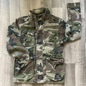 Camouflage parkajacka från C Division - Säljer en grön och brun camouflage parkajacka från C Division med stl M. Jackan har flera stora fickor framtill, hög krage och dragkedja. Perfekt för dig som gillar militärstil och vill sticka ut lite extra.
