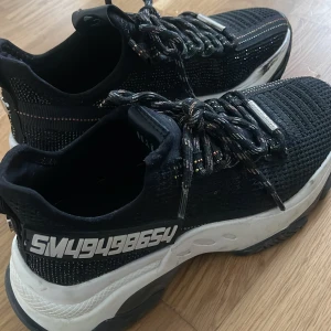 Chunky svarta sneakers med glitterdetaljer - Svarta chunky sneakers med vit och svart sula, glittriga detaljer i tyget och coola snören med färginslag. Sidan har en stor vit textdekor. Ovandelen är stickad och ger en sportig vibe, perfekt för dig som gillar statement-skor.