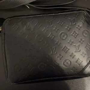 Svart axelväska  - Säljer en svart axelväska med klassiskt monogrammönster i präglat skinn. Väskan har en justerbar axelrem och stilren design som passar perfekt till streetwear eller en mer dressad look.