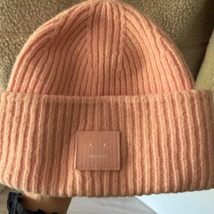 Rosa  mössa Acne Studios - Superfin rosa mössa från Acne Studios med ribbstickad struktur och det ikoniska face-patch framtill. Tillverkad i mjuk ull som håller dig varm och snygg. Perfekt accessoar för att lyfta din vinterlook.