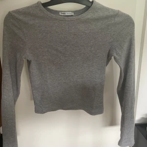 Grå långärmad topp från Bershka - En basic grå långärmad topp från Bershka i mjuk bomullsmix. Modellen är croppad med rund halsringning och har en slim passform som sitter snyggt på kroppen. Perfekt att styla med jeans eller kjol för en clean look.