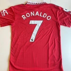 Manchester United Ronaldo 7 Adidas tröja - Manchester Uniteds officiella matchtröja replika med Ronaldo 7-tryck. Liten spricka i ena trycket på armen enligt bild. Röd med svarta och vita detaljer, vit krage med röd dekor, klubbmärke och Adidas-logga på bröstet. Kortärmad och tillverkad i lätt, ventilerande material perfekt för fotboll. Endast handtvätt gäller.