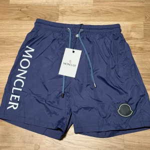 Blå Moncler badshorts med snörning - Snygga marinblå badshorts från Moncler med vit logga längs benet och klassisk badge framtill. Shortsen har elastisk midja med snörning och innerbyxa i mesh för extra komfort. Perfekta för stranden eller poolen, riktigt clean look.