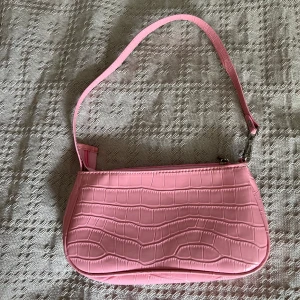 Rosa axelväska i croco-imitation - Säljer en söt rosa axelväska i syntetmaterial med croco-mönstrad yta. Väskan har en smal axelrem och stängs med dragkedja. Perfekt storlek för det viktigaste och ger en poppig vibe till din outfit.