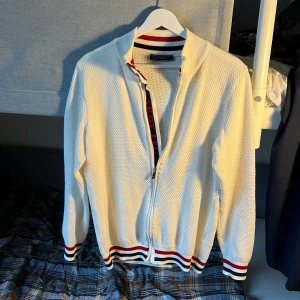 Vit cardigan jacka från Felix Hardy - Snygg vit cardigan jacka från Felix Hardy med strukturstickat tyg, dragkedja framtill och ribbade muddar med röd, blå och vit rand. Jackan har lång ärm och detaljer med logga längs dragkedjan. Perfekt för dig som gillar stilren look.